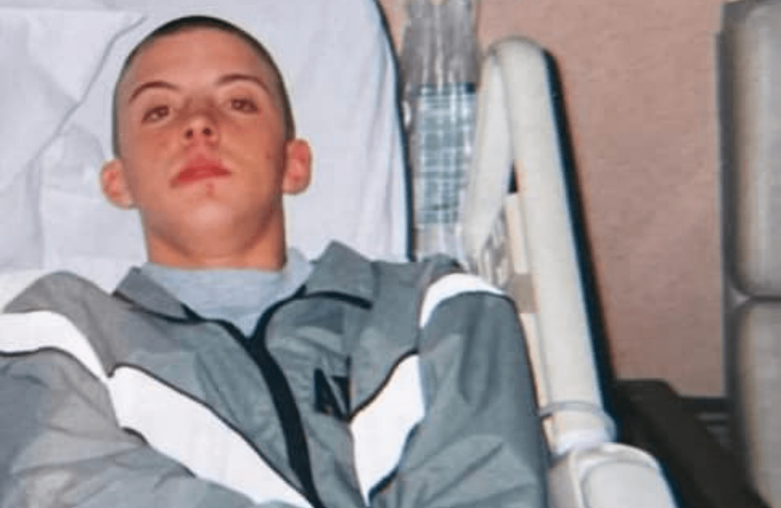 Teenage boy in grey jacket lying in hospital bed with IV line, looking into camera