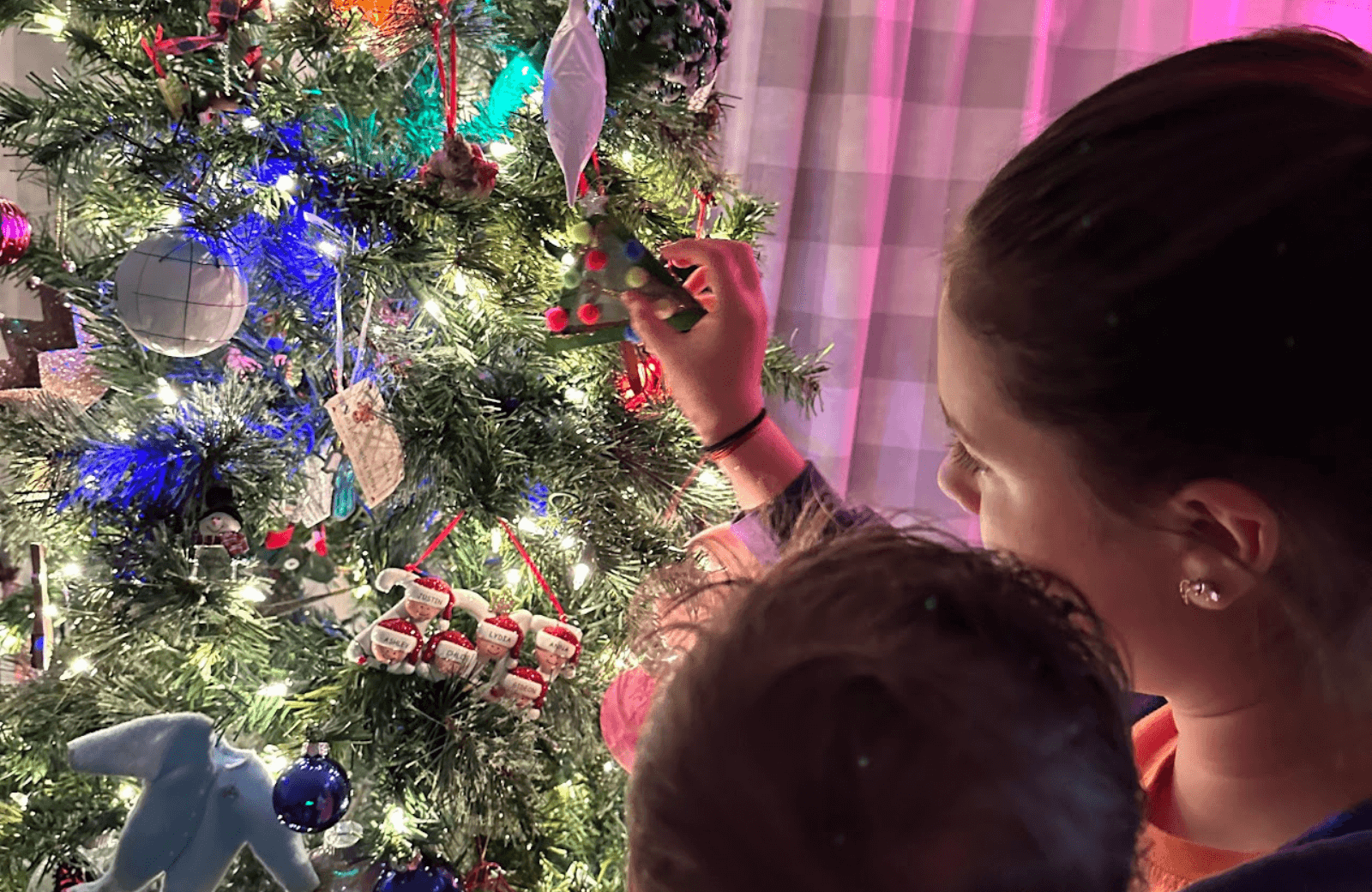 Young girl decorating a Christmas tree while holding a baby, warm glow of lights in a living room